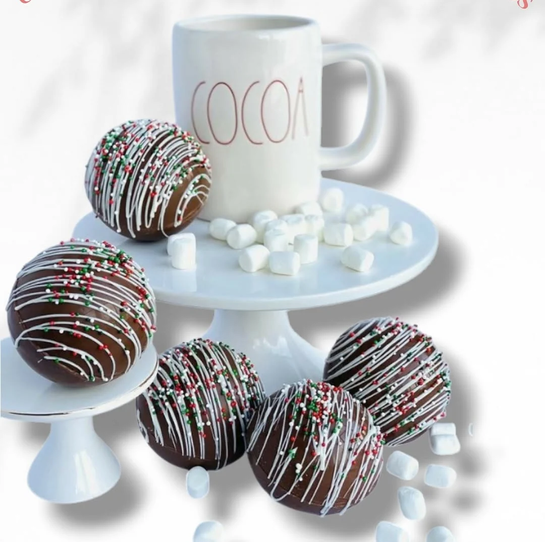 Five Christmas cocoa bombs decorated with white drizzle and sprinkles, beside a cocoa mug and marshmallows, Detalles de Amor Medellín